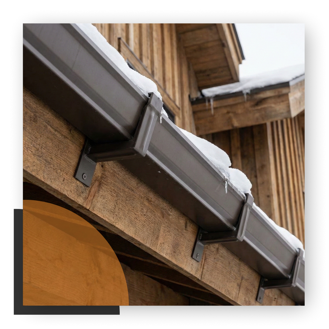 Dark brown seamless gutters along the eave of a wood-sided home with snow on the roof above